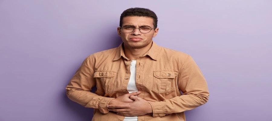Imagem do post Homem com expressão de dor segurando o abdômen, aparentando desconforto estomacal ou cólica, em frente a um fundo lilás.