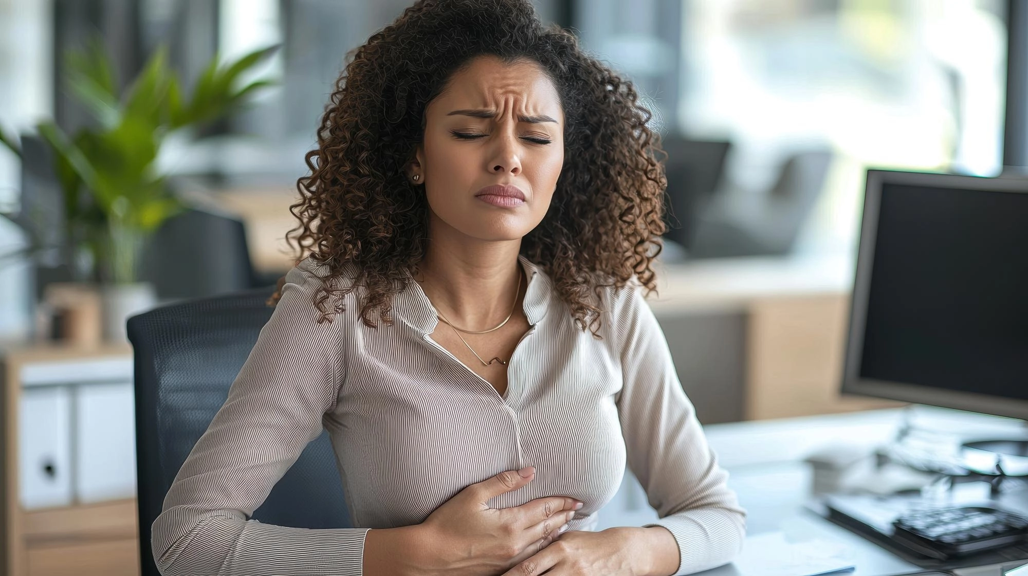 Imagem do post Mulher no escritório com as mãos sobre a barriga e semblante de dor por causa de uma dor no estômago que não passa