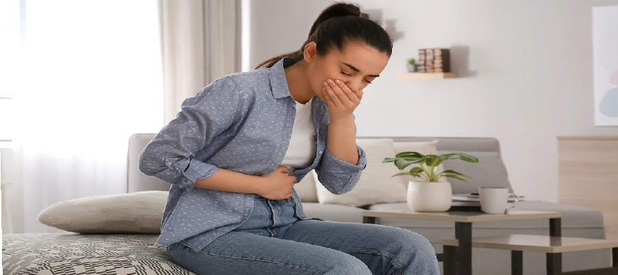 Imagem do post Mulher com expressão de náusea sentada na cama, segurando o estômago com uma mão e cobrindo a boca com a outra, indicando mal-estar gastrointestinal em ambiente doméstico.