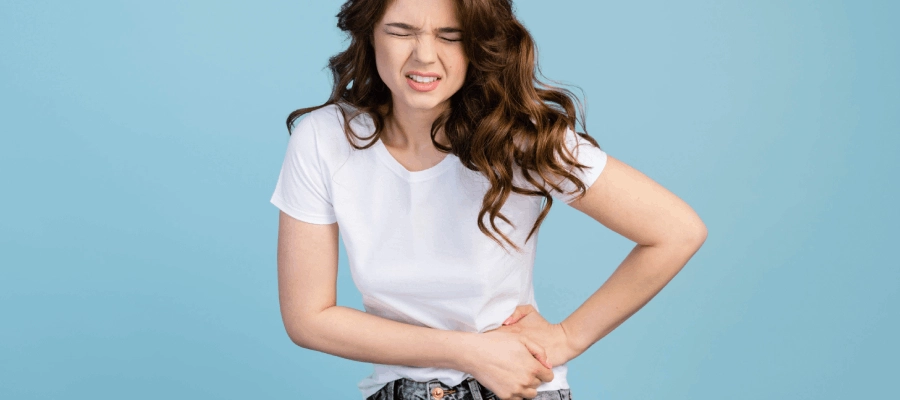 Imagem do post Mulher jovem em pé segurando a lateral do abdômen com expressão de dor e de pontada no lado esquerdo da barriga.