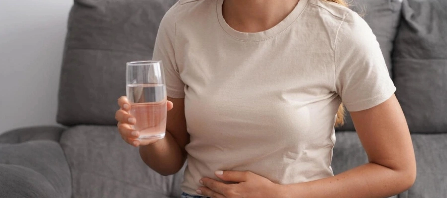 Imagem do post Mulher jovem sentada no sofá de casa, sentindo dores abdominais, segura um copo de água para tomar remédio.