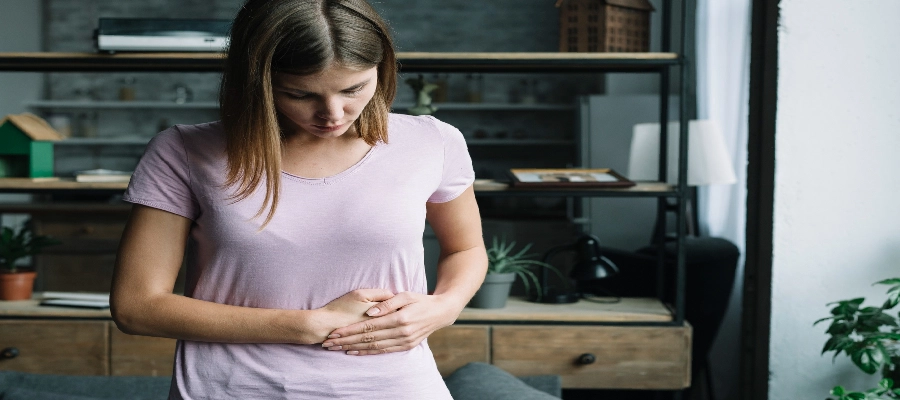 Imagem do post Mulher com expressão de dor olhando para o abdômen e segurando a barriga, indicando cólica ou dor abdominal em ambiente doméstico.