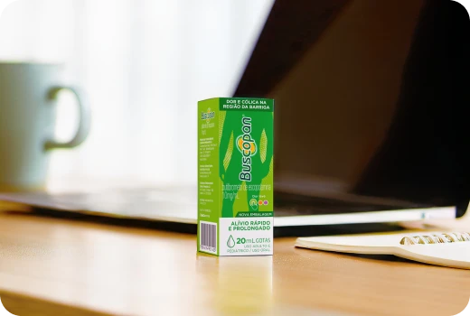 Embalagem do produto Buscopan Gotas em cima de uma mesa, ao lado de um notebook e uma caneca.